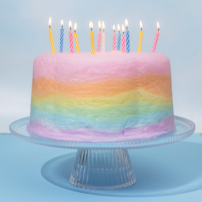rainbow cotton candy cake with several lit candles on top displayed on a clear glass cake stand in front of a light blue background