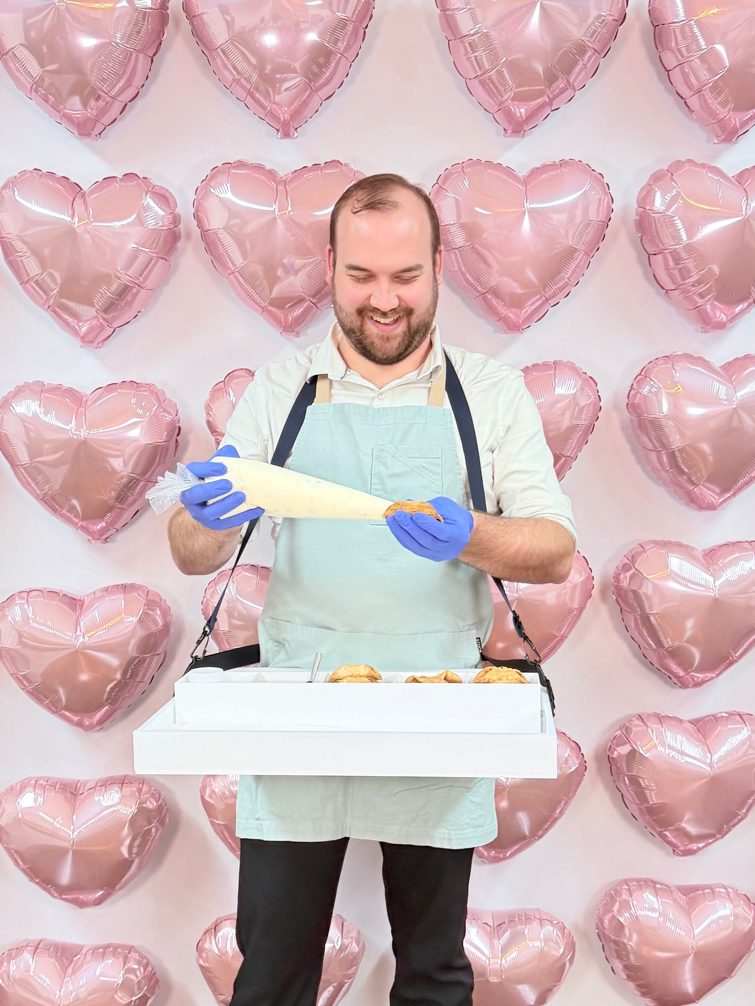 man adding cream to a cannoli with a serving tray in front of a decorative heart balloon wall