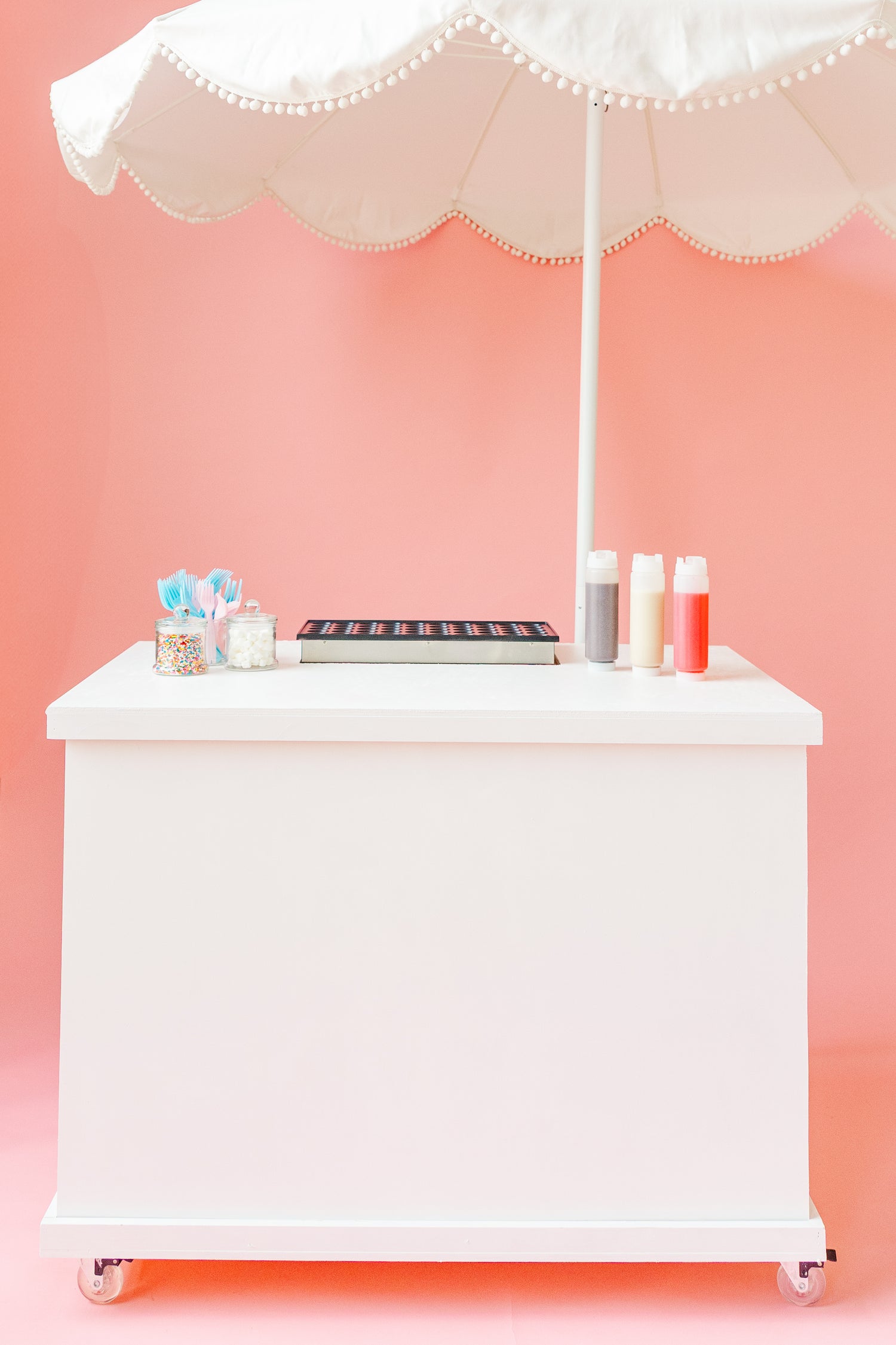 white cart with pancake griddle, sprinkle containers, and three squeeze bottles of syrups on top with a white umbrella in front of a pink background