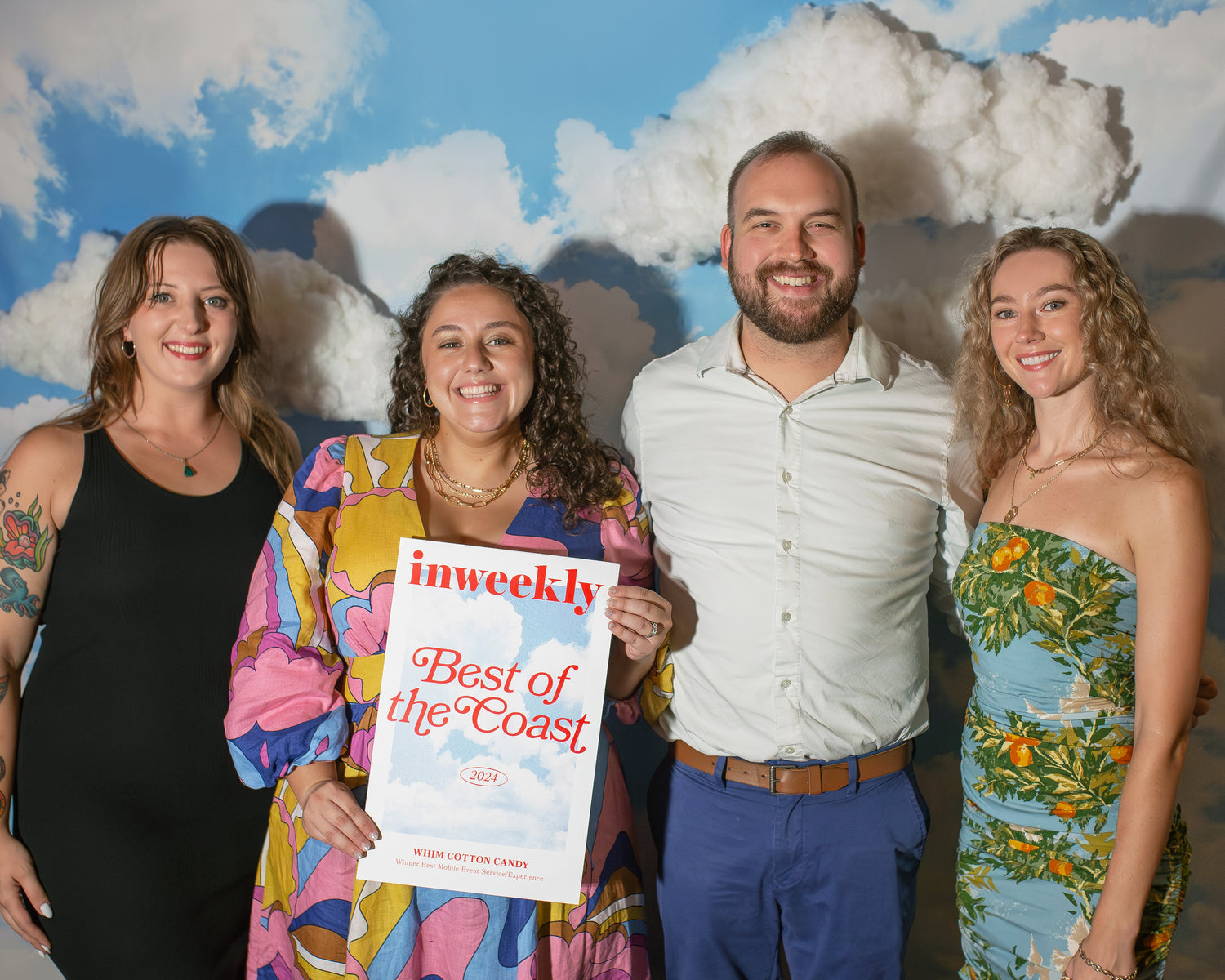 Four members of the Whim team posing with a 'Best of the Coast' award against a cloud-patterned backdrop